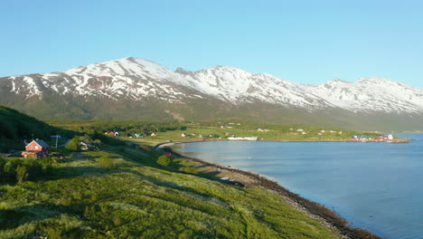 Lufttief,-Drohnenschuss-über-Häuser-In-Einem-Kleinen-Dorf-An-Der-Küste-Der-Barentssee,-Schneebedeckte-Berge-Im-Hintergrund,-Sonniger-Tag,-In-Troms,-Nordland,-Nordnorwegen