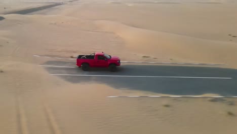 Bright-red-truck-driving-onto-a-single-road-with-sand-spilling-over-from-desert,-drone-tracking-from-the-side