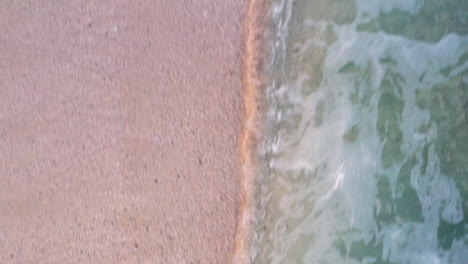 Aerial-ascending-shot-of-a-sandy-beach,-starting-from-a-close-up-of-the-waves-hitting-the-sand,-then-ascending-to-reveal-the-beach