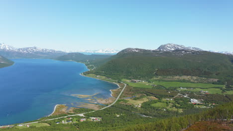 Aerial-view-over-a-mountain,-overlooking-a-village-in-a-valley,-in-the-fjords,-on-the-coastline-of-the-Arctic-ocean,-sunny,-summer-day,-in-Rotsund,-Troms,-Nordland,-Norway---pan,-drone-shot