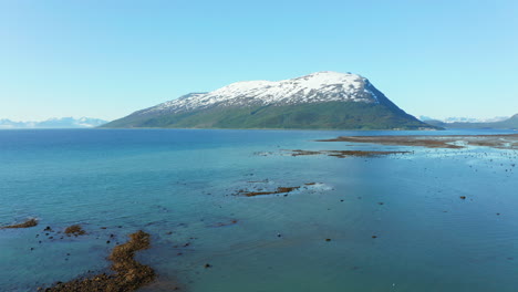 Aerial,-rising,-drone-shot-of-tranquil,-blue-sea-and-a-snowy-mountain,-sunny,-summer-day,-in-the-Lyngen-alps,-Troms,-Nordland,-North-Norway