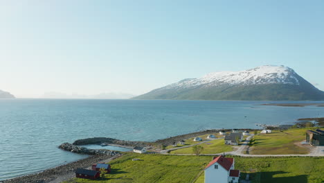 Aerial-view-over-houses-and-a-harbor,-in-a-small-village-on-the-coast-of-the-Barents-sea,-snowy-alpine-in-the-background,-sunny-day,-in-Troms,-Nordland,-North-Norway---low,-dolly,-drone-shot