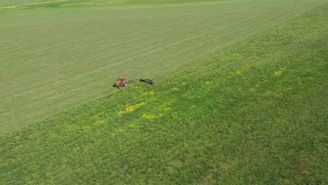 Haymaking-in-sunny-green-field-with-tractor-and-tow-behind-mower,-long-drone-shot