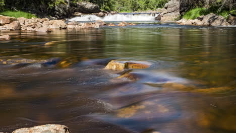 Time-lapse-of-shallow-river-flowing-swiftly-small-waterfall-in-background