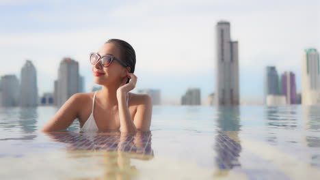 While-lounging-in-a-rooftop-infinity-pool-backed-by-the-modern-skyline-of-the-city-of-Bangkok,-Thailand-a-beautiful,-young-woman-turns-her-face-toward-the-sun