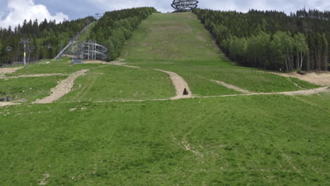 Tracking-aerial-4k-shot-of-a-four-wheeler-driving-up-a-green-slope-in-the-mountains-of-Dolní-Morava,-Czechia,-heading-towards-The-Sky-Walk-attraction-between-forests
