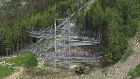 Rotating-aerial-4k-shot-of-a-big-loop-of-an-extreme-outdoor-roller-coaster-ride-in-the-mountains-of-Dolní-Morava,-Czech-Republic,-with-trees-and-a-cableway-in-the-background