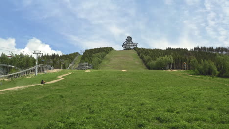 Zooming-aerial-4k-shot-of-a-four-wheeler-driving-up-a-green-slope-heading-towards-The-Sky-Walk-attraction-in-Dolní-Morava,-Czechia-with-a-cableway-and-forests-around