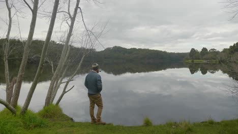 A-wide-shot-of-a-man-standing-by-a-lake-and-reflecting-while-smoking-a-tobacco-pipe