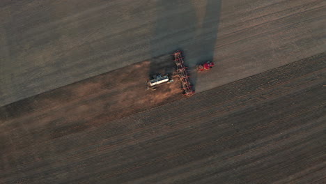 Stunning-view-of-seeding-farm-tractor-machine-traveling-on-flat-dirt-and-dusty-farmland-rows-at-sunset,-Saskatchewan,-Canada,-directly-above-aerial-static