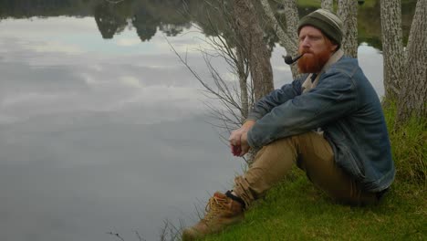 A-mid-shot-of-a-bearded-ginger-mountain-man-smoking-a-tobacco-pipe-while-sitting-by-a-calm-lake
