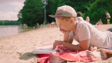 Cute-baby-boy-with-a-cap-lying-on-a-sandy-lakeshore,-reading-the-book,-and-having-his-tropical-vacation