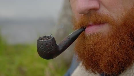 A-close-up-shot-of-a-ginger-bearded-man-smoking-a-tobacco-pipe-and-blowing-out-smoke