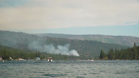 California-Wildfire,-mountain-burns-on-edge-of-lake,-helicopter-drops-water-on-smoke-plume
