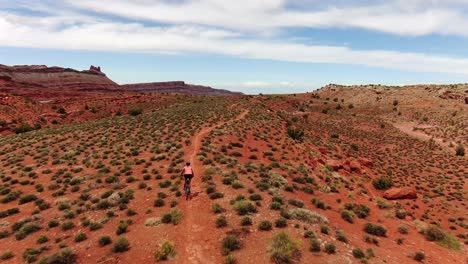 Aerial-follow-shot-behind-woman-mountain-biking-on-flat-trail-with-red-desert-sandstone