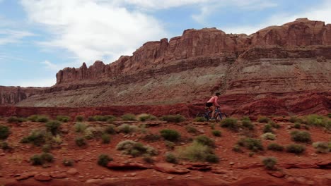 Aerial-shot-following-woman-riding-mountain-bike-in-red-Moab-desert-with-mountains