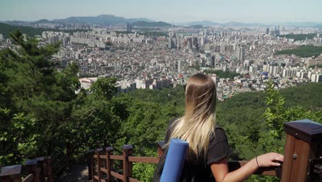 Blondes-Mädchen,-Das-Sich-An-Den-Zaun-Lehnt-Und-Auf-Die-Schöne-Stadt-Des-Seocho-gu-bezirks-Mit-Hochhäusern-Vom-Gwanaksan-bergpfad-In-Seoul,-Südkorea,-Blickt
