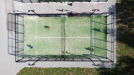Four-fit-healthy-young-people-playing-paddle-tennis-at-a-green-outside-outdoors-paddle-court-tennis-balls-hitting-racket-sport-being-sweaty-fitness-activity-from-above-playing-together-as-team-friends