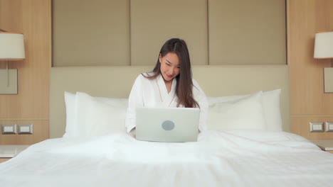 While-sitting-in-a-bathrobe-in-a-kingsize-hotel-bed-a-pretty,-young-woman-works-on-her-laptop