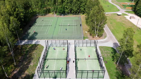 Aerial-tilting-down-on-four-green-outdoor-paddle-tennis-courts-filled-with-athletic-fitness-sports-people-training-and-playing-racket-sports-at-golf-resort-near-sea-side-at-summer-tennis-paddle-camp