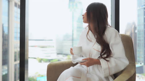 Pretty-Asian-Female-Drinking-Morning-Coffe-in-Bathrobe-by-Window-of-Family-Home