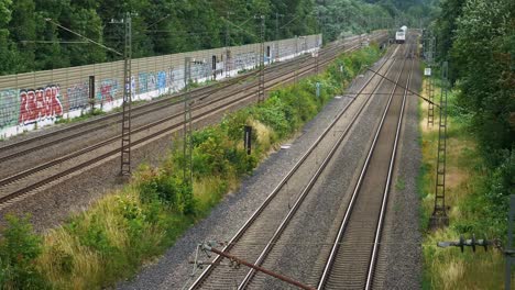 A-metallic-train-approaches-on-a-pair-of-tracks-in-a-rural-area-with-tall-bushes-and-trees-on-the-side