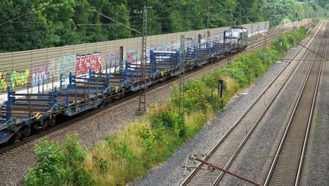 A-large-shipping-train-with-empty-flatbeds-behind-it-goes-down-the-line-of-railroad-bordered-by-a-graffiti-covered-wall