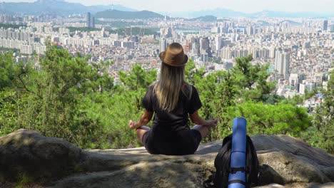 Girl-Sitting-On-The-Rock-And-Practicing-A-Yoga-Lotus-Pose-Against-The-Skyline-Of-Seocho-gu-District-From-The-Gwanaksan-Mountain-In-Seoul,-South-Korea