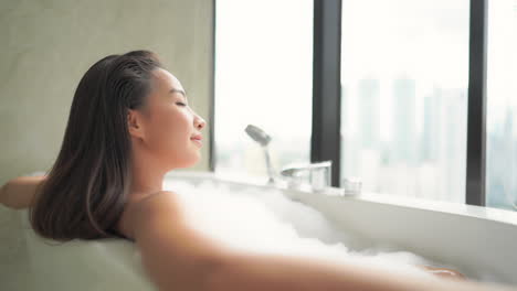 Pretty-Asian-Woman-Enjoying-in-Foamy-Bathtub-With-Windows-View-on-Modern-City,-Close-Up