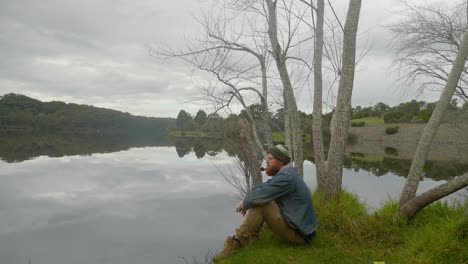 A-wide-shot-of-a-bearded-ginger-man-sitting-by-a-lake-and-smoking-a-tobacco-pipe