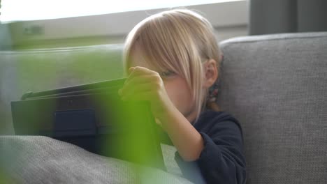 Little-girl-uses-tablet-at-home-on-couch,-slider-shot