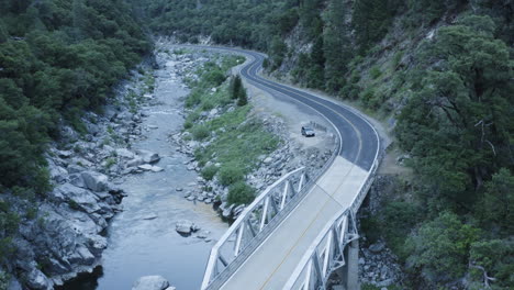 Small-bridge-crosses-over-flowing-water-to-connect-two-sides-of-valley,-aerial
