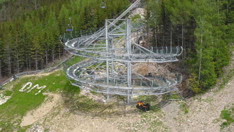 Rotating-aerial-4k-shot-of-a-cart-with-people-spiralling-down-a-big-loop-of-an-extreme-outdoor-roller-coaster-in-the-mountains-of-Dolní-Morava,-Czech-Republic-with-trees-around
