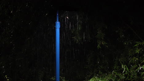 Summertime-torrential-rain-pours-down-past-light-cast-from-a-blue-lamp-post-surrounded-by-green-foliage-at-night-as-insects-dance-around-the-light-source