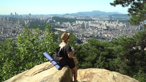 Girl-Sitting-On-The-Boulder-And-Drinking-After-A-Long-Hike-And-Looking-Towards-The-Downtown-Skyline-Of-Seocho-gu-District-From-The-Gwanaksan-Mountain-In-Seoul,-South-Korea