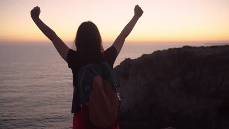 Rear-view-of-young-girl-raising-hands-in-achievement-at-dawn-on-top-of-cliff,-slow-motion