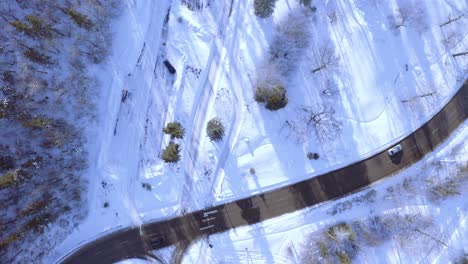 Aerial-hold-over-birds-eye-view-curvy-hiyway-freeway-road-in-the-winter-central-to-a-snow-covered-park-forest-with-light-vehicle-car-traffic-passing-by-as-the-sun-shadows-the-bare-trees