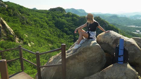 Girl-Sitting-On-The-Boulders-With-Green-Forest-And-City-Skyline-On-The-Background-From-The-Gwanaksan-Mountain-In-Seoul,-South-Korea