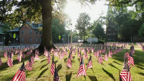 Establishing-shot-of-small-town-in-United-States-with-lawn-decorated-with-American-flags-during-dramatic-morning-sunlight-through-trees,-church-in-distance