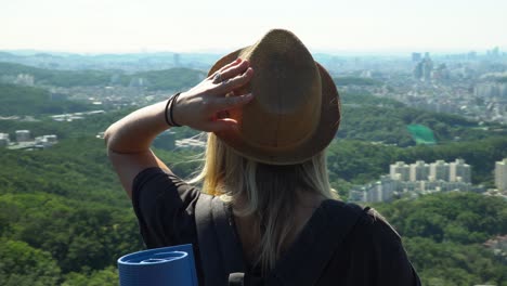 Girl-In-Hat-And-Sunglasses-With-Flowing-Hair-Leaning-On-The-Fence-And-Enjoying-The-Downtown-Skyline-From-The-Gwanaksan-Mountain-During-Summertime-In-Seoul,-South-Korea