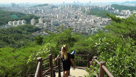 Young-Caucasian-Girl-Climbing-Up-On-The-Trail-While-Holding-On-The-Wooden-Railings-With-A-City-View-From-The-Gwanaksan-Mountain-On-A-Sunny-day-In-Seoul,-South-Korea