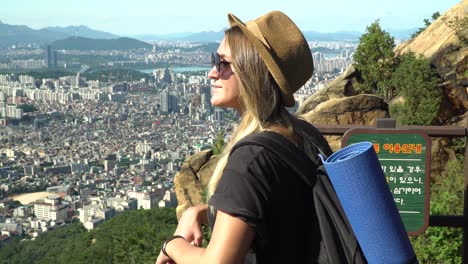 Stylish-Blonde-Girl-With-Hat-And-Sunglasses-Leaning-On-The-Fence-And-Admiring-The-Beautiful-City-Of-Seocho-gu-From-The-Gwanaksan-Trail-On-A-Sunny-Day-In-Seoul,-South-Korea