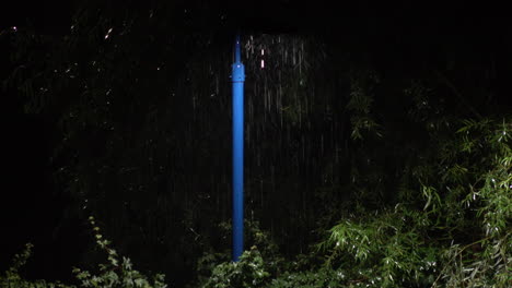 Im-Sommer-Ergießt-Sich-Sintflutartiger-Regen-über-Das-Licht,-Das-Von-Einem-Blauen-Laternenpfahl-Geworfen-Wird,-Der-Nachts-Von-Grünem-Laub-Umgeben-Ist,-Während-Insekten-Um-Die-Lichtquelle-Tanzen