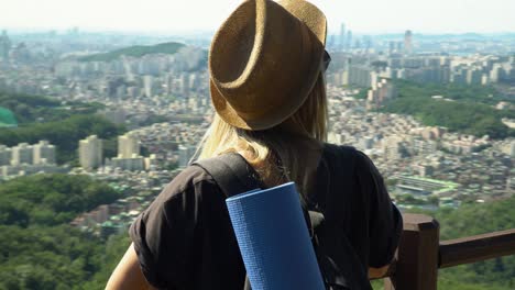 Young-Caucasian-Girl-In-Brown-Hat-Leaning-On-The-Railings-And-Looking-Away-At-The-Beautiful-City-Of-Seocho-gu-District-From-The-Gwanaksan-Mountain-In-Seoul,-South-Korea