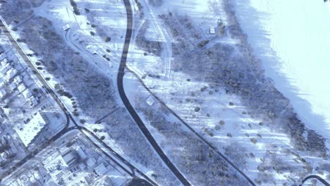 Birds-eye-view-aerial-pan-left-of-a-highway-and-an-ice-snow-covered-river-next-to-park,-next-to-town