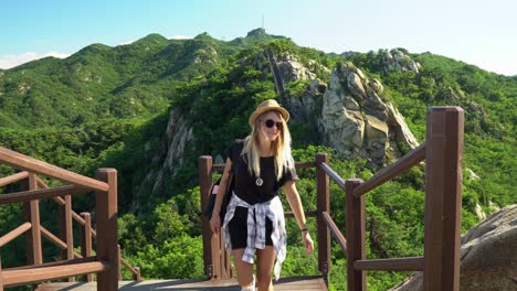 Cute-blond-backpacker-girl-on-a-hike-to-summit-enjoying-sunny-landscape,-holding-railing-and-walking-up-stairs-to-continue-trekking