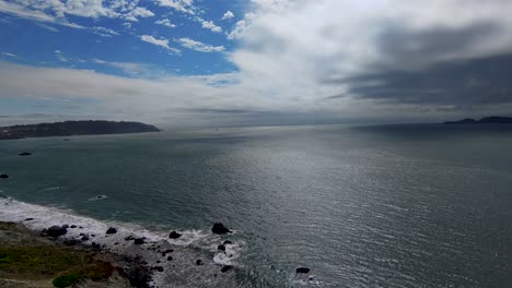 Ruhiges-Meerblickpanorama-Mit-Wellen,-Die-Die-Felsen-An-Land-Treffen,-Und-Blauer-Bewölkter-Himmelhintergrund