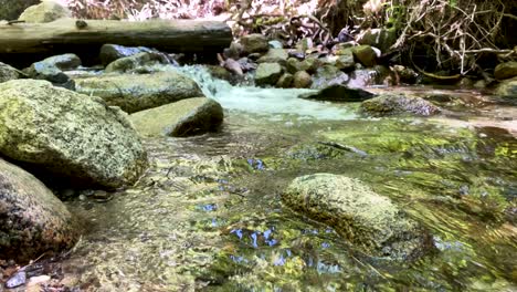 Smooth-slow-motion-shot-floating-over-flowing-creek,-moving-towards-waterfall-in-mountain-stream,-with-large-river-rocks-in-foreground-and-pebbles-under-the-clear-water,-green-from-trees-overhead
