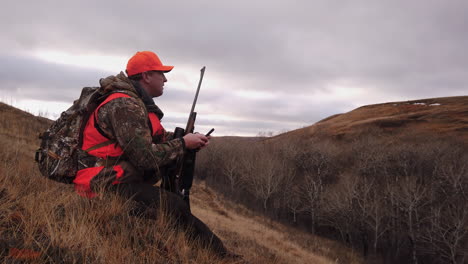 Jäger-Mit-Einem-Gewehr,-Der-Auf-Einem-Grasbewachsenen-Hügel-Sitzt-Und-Ein-Handy-Suchsignal-In-Einem-Jagdgebiet-In-Saskatchewan,-Kanada-Hält---Mittlerer-Schuss