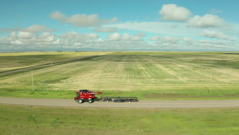 Modern-red-combine-harvester-versatile-machines-drive-on-dirt-roads-by-expansive-flat-green-farmland-of-crops-on-sunny-day,-Saskatchewan,-Canada,-above-aerial-sideways